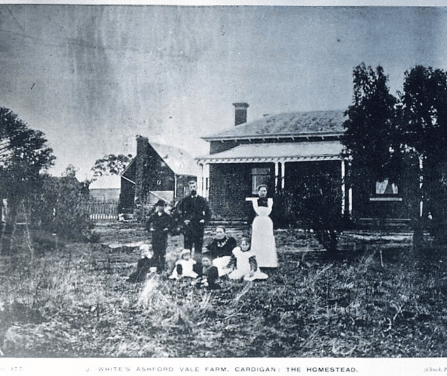 An old, grainy black and white photo shows two adults and one child standing, and one adult sitting with four children in front of a brink house with a veranda.