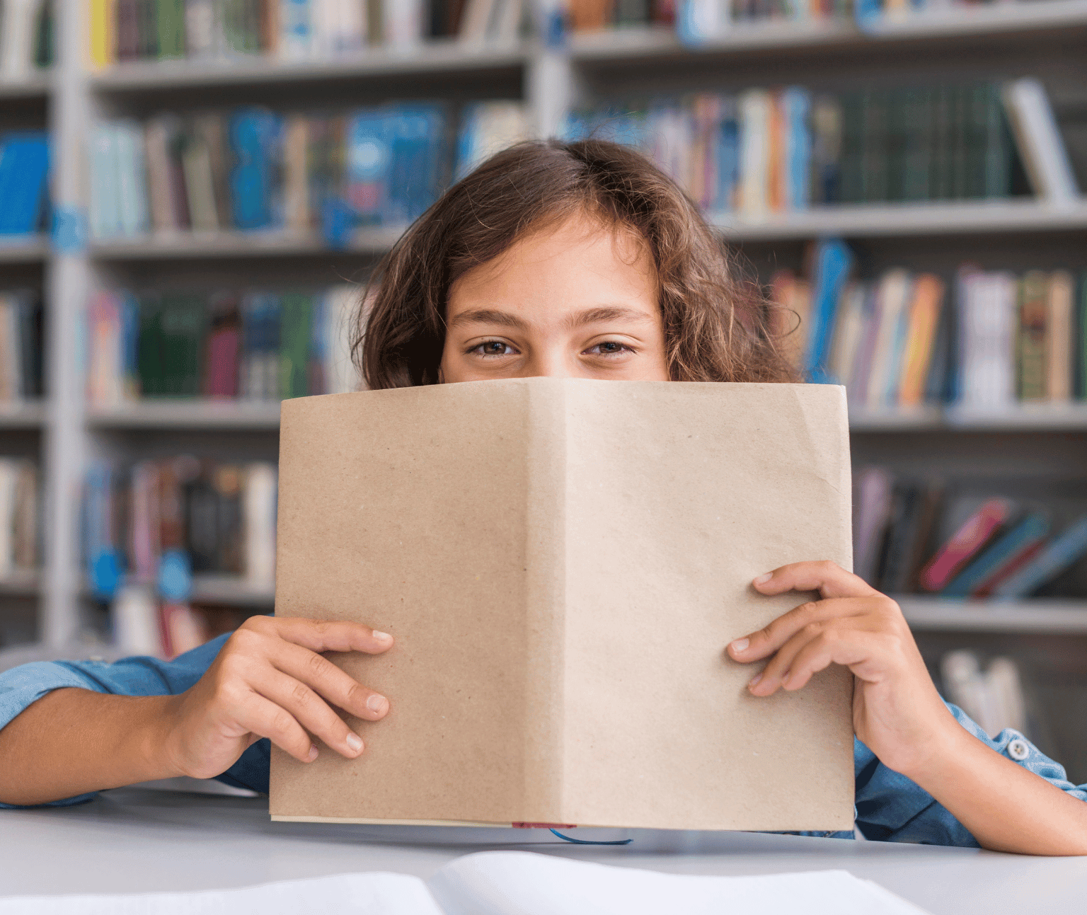 A child with brown hair and brown eyes peers over the top of an open book that is covered in light brown paper.