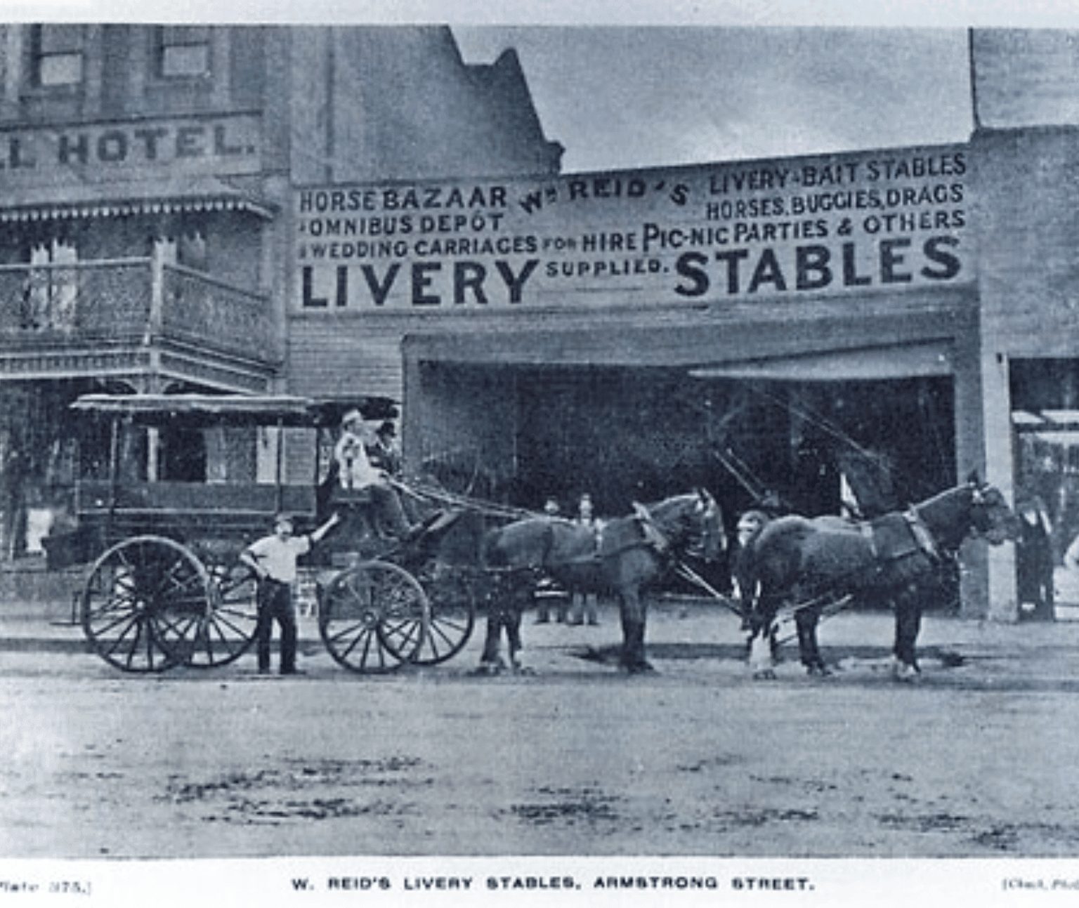 An old black and white photo shows a horse drawn coach pulled by four horses. Two people sit in the front of the coach and one person is leaning against it. Behind them is a building the words 'Livery Stable' painted on above a wide entrance.