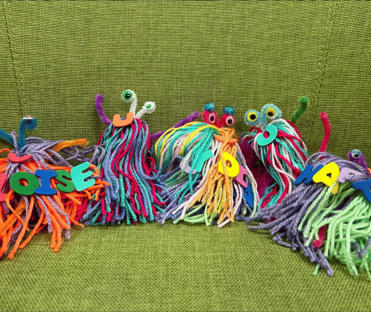 Four pom pom monsters made from colourful yarn have two googly eyes each. The are sitting against green fabric.