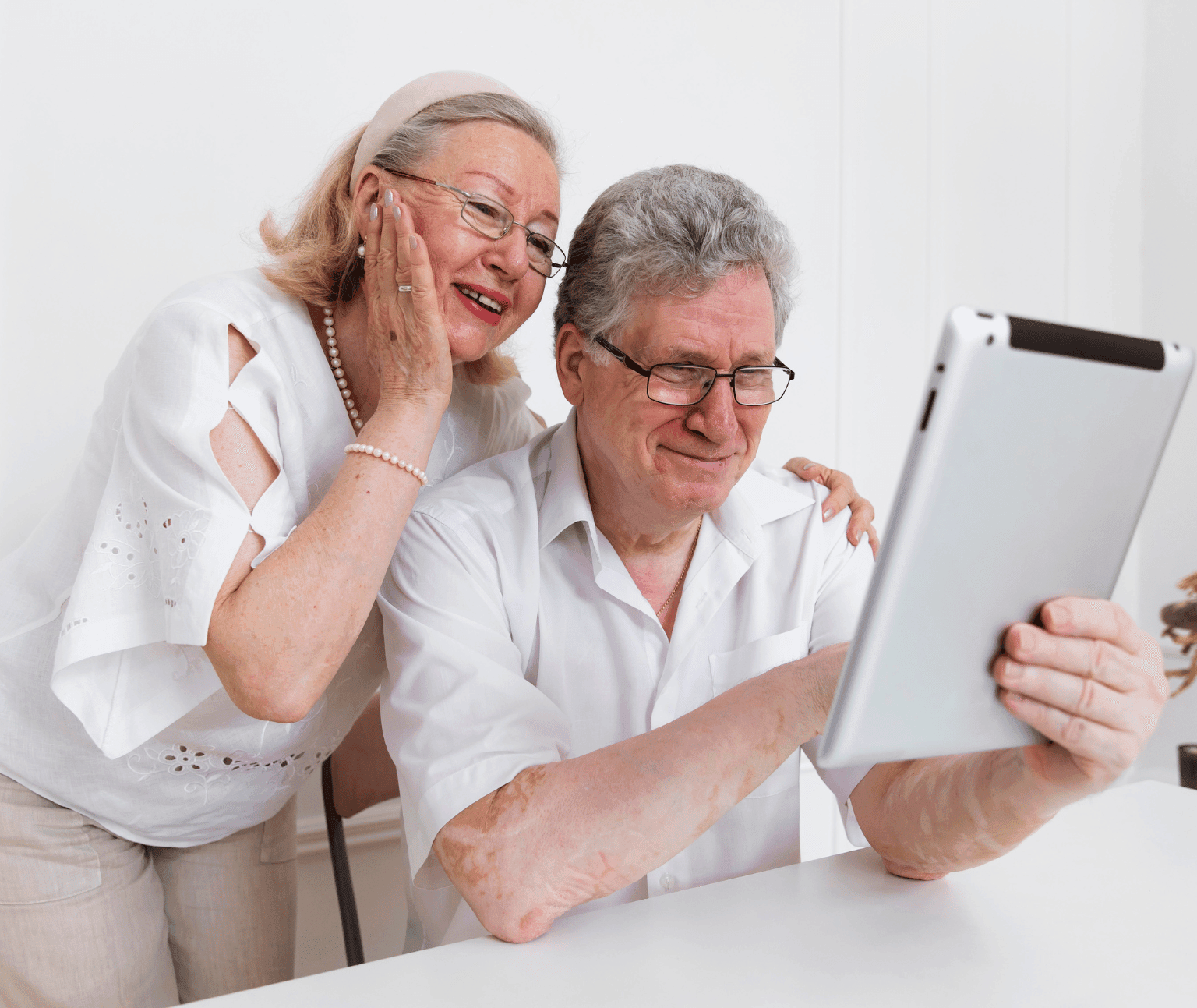 An older female looks over an older male's shoulder. they are both smiling and looking at the screen of an iPad.