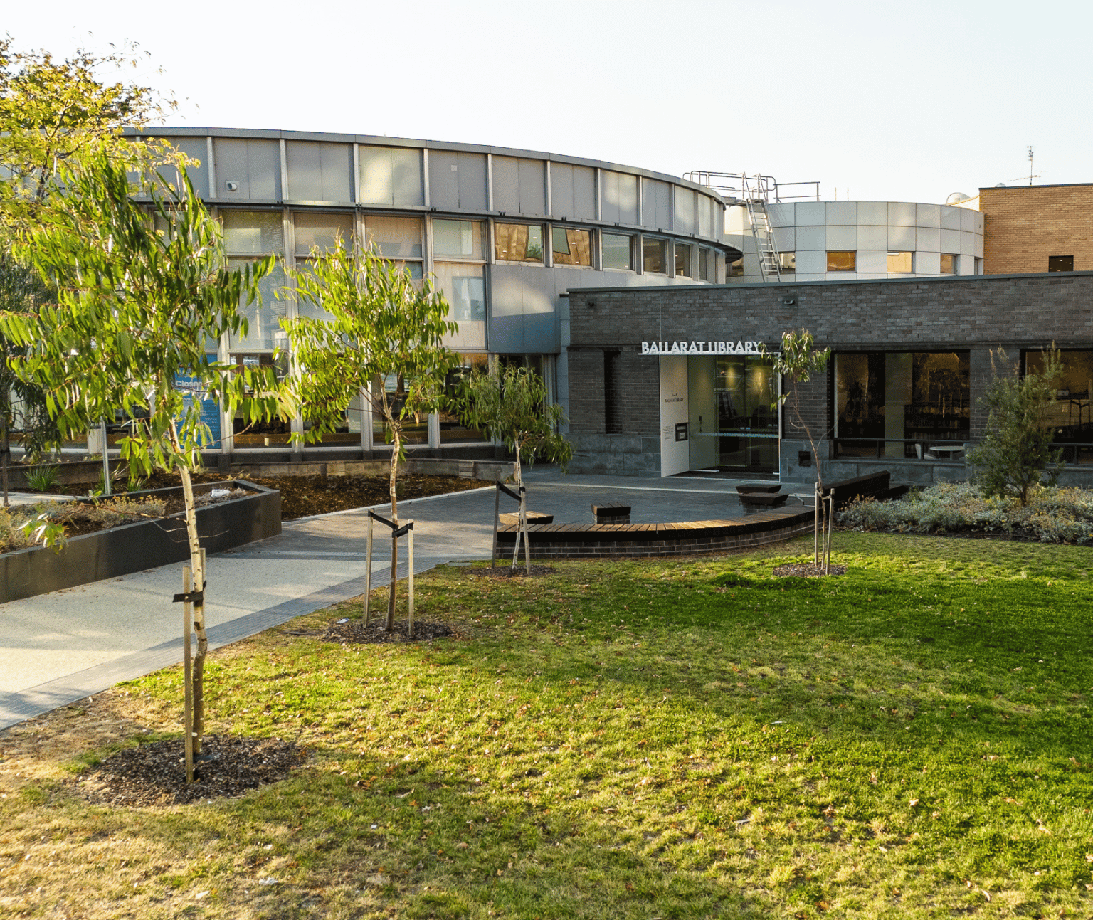 An external view of the Ballarat Library, showing the new entrance with library sign over the door. There is grass, small gum trees and garden beds. The library has large windows and a dark grey brick.