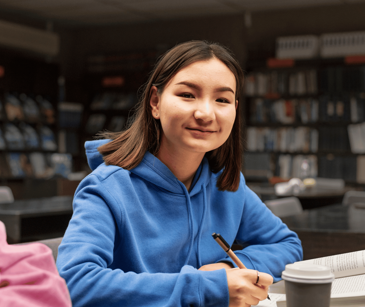 A teenage girl with brown hair, wearing a blue jacket sits. there are shelves with books on them in the background.