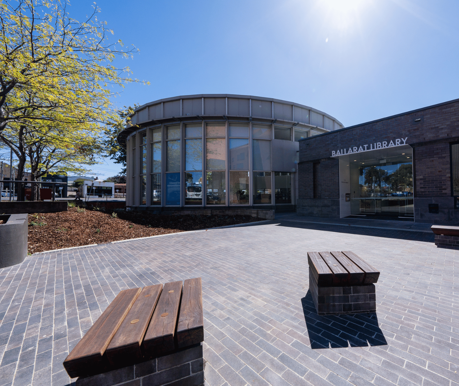 An external photo of the Ballarat Library. There are large glass automatic sliding doors, as well as a circular part of the building which has large windows all the way arounds. There is a new garden bed with small plants and a paved walkway.