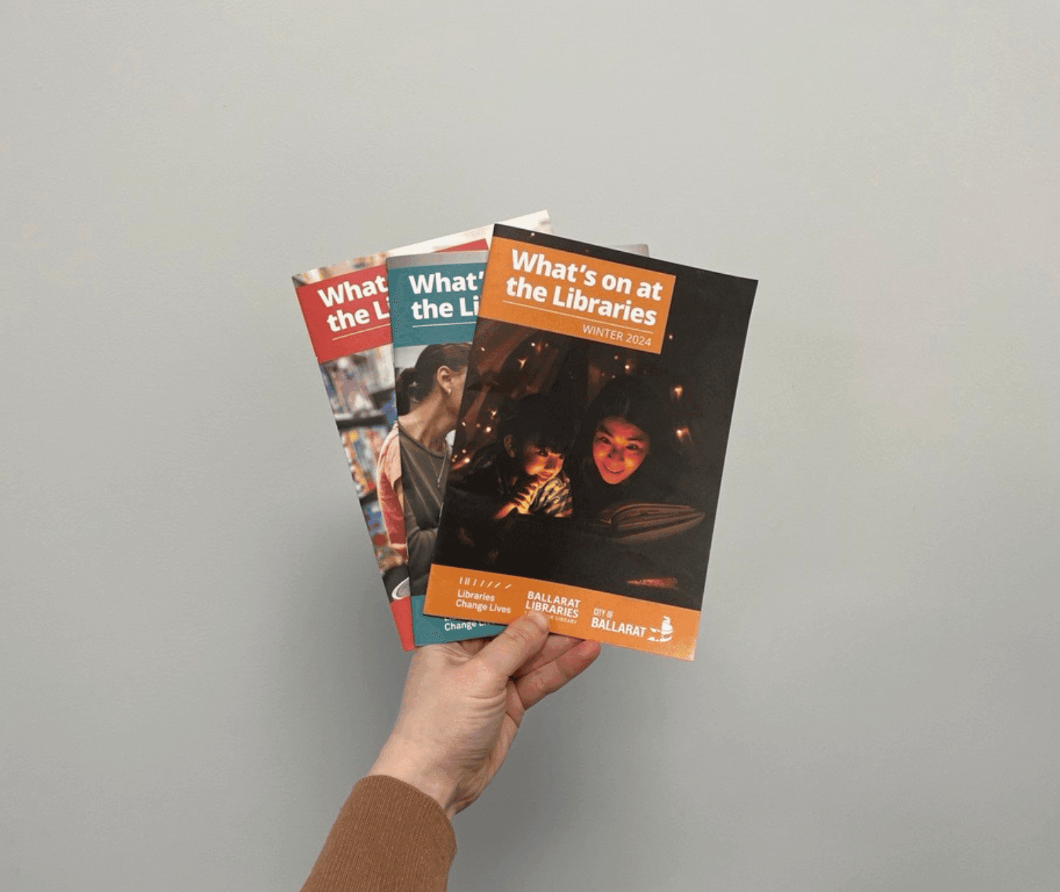 A hand holds three Ballarat Libraries What's On brochures. The front brochure has the text 'What's on at the Libraries Winter 2024' and an adult and child looking at a book in dim light with fairy lights above them.
