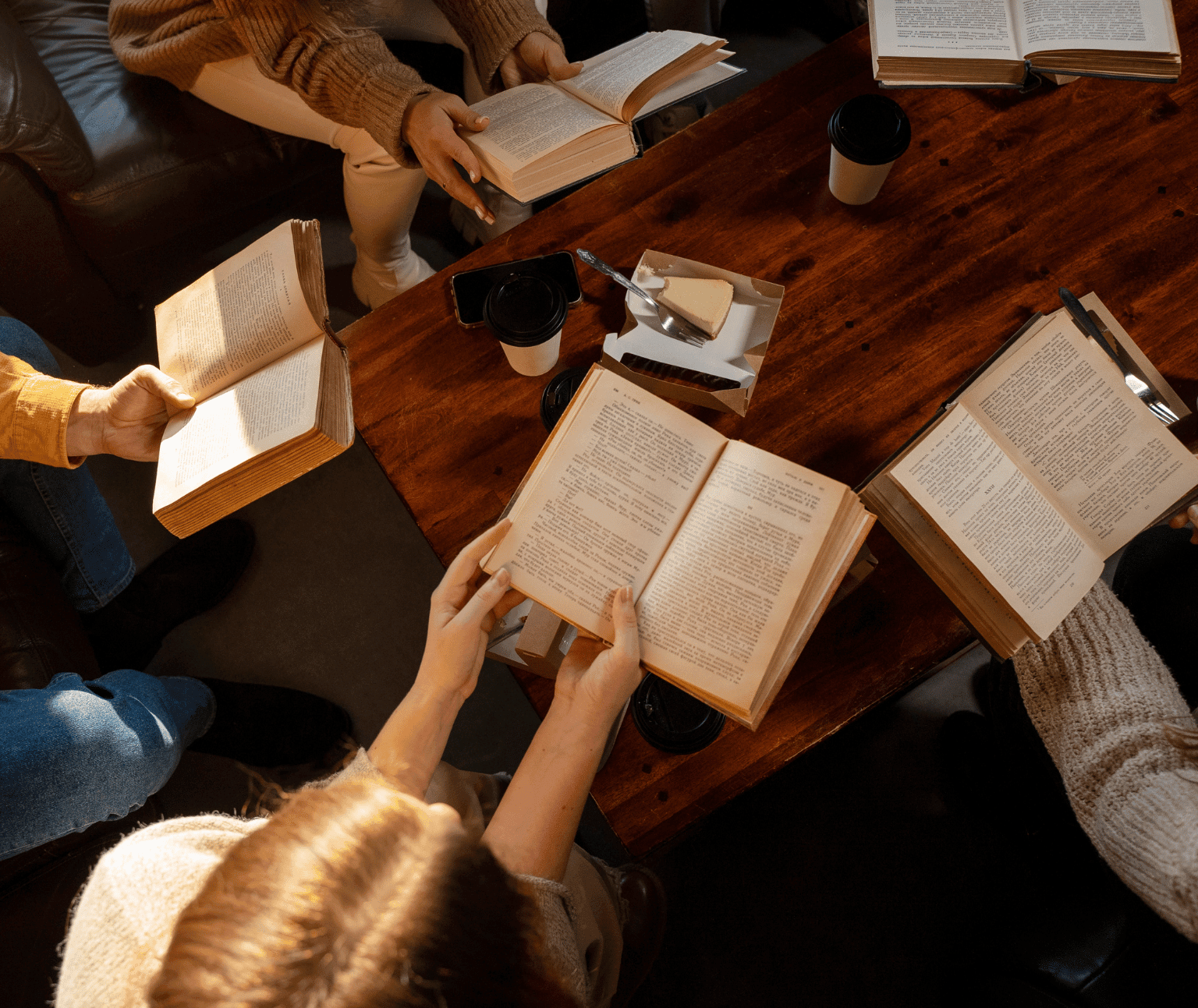 Three people sit around a table, each holding a book that is open. A take away coffee cup, slice of cake and an iPhone are on the table.