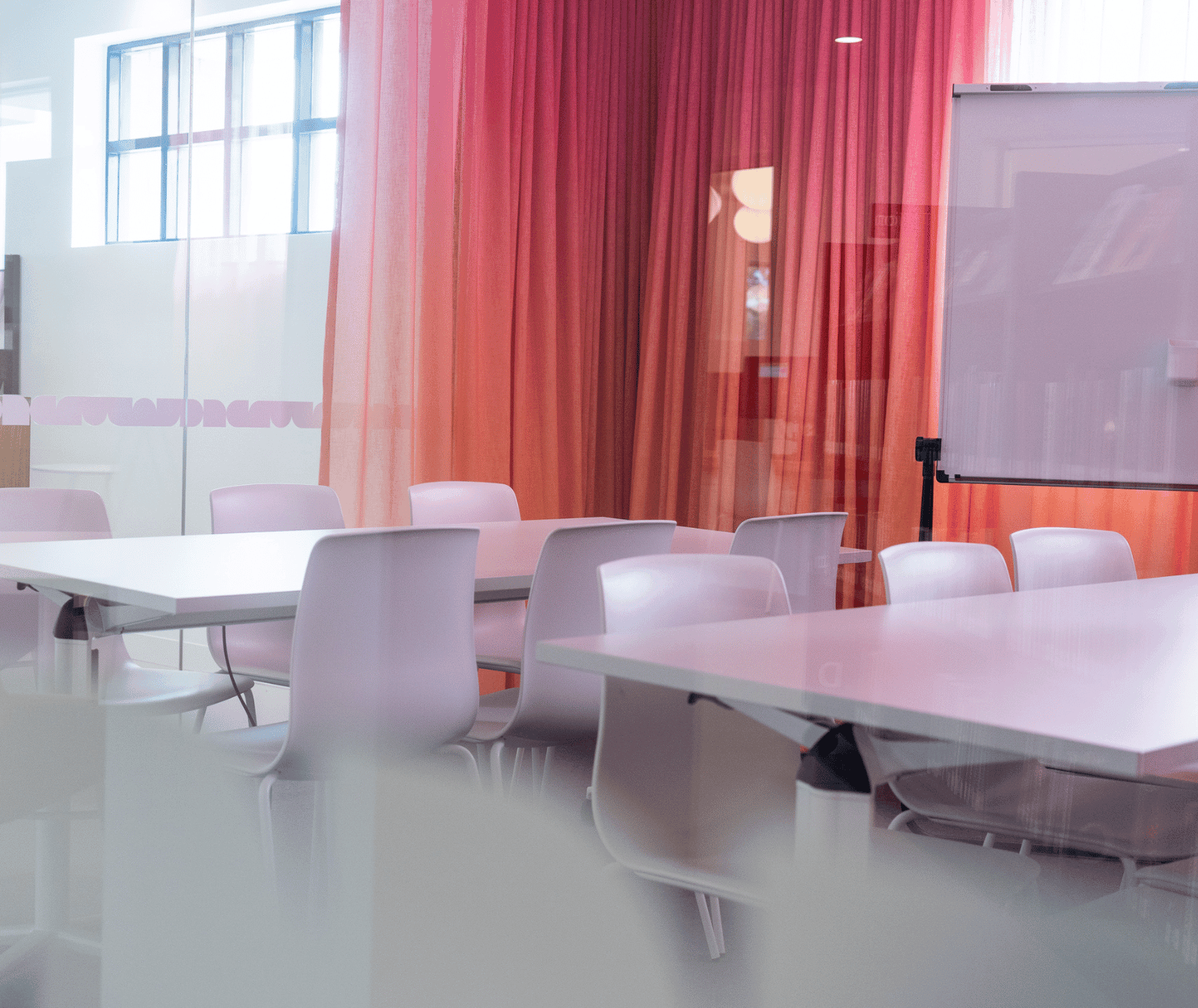 A Ballarat Library meeting room with two white, rectangular tables with chairs around them. Pink and orange curtains have a whiteboard sitting in front of them.