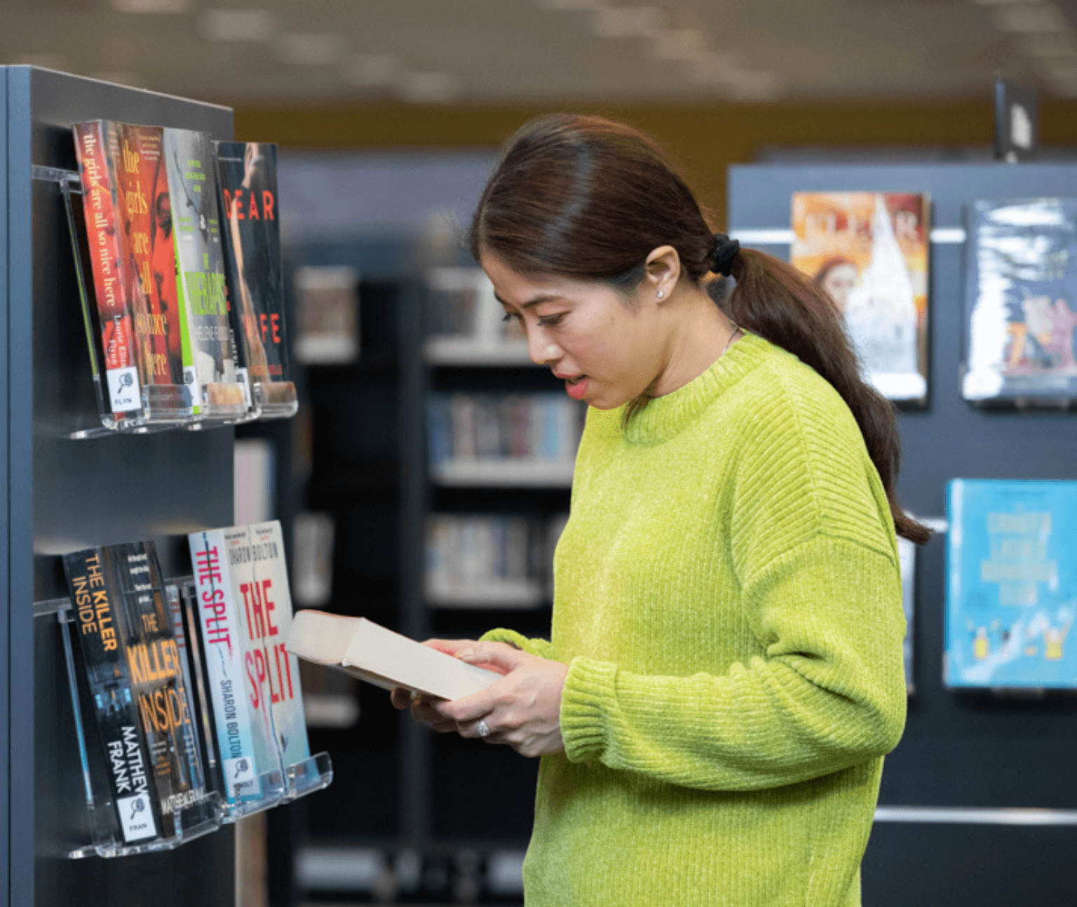 A female wearing a lime green jumper reads the blurb of a book. There are shelves of books behind her.
