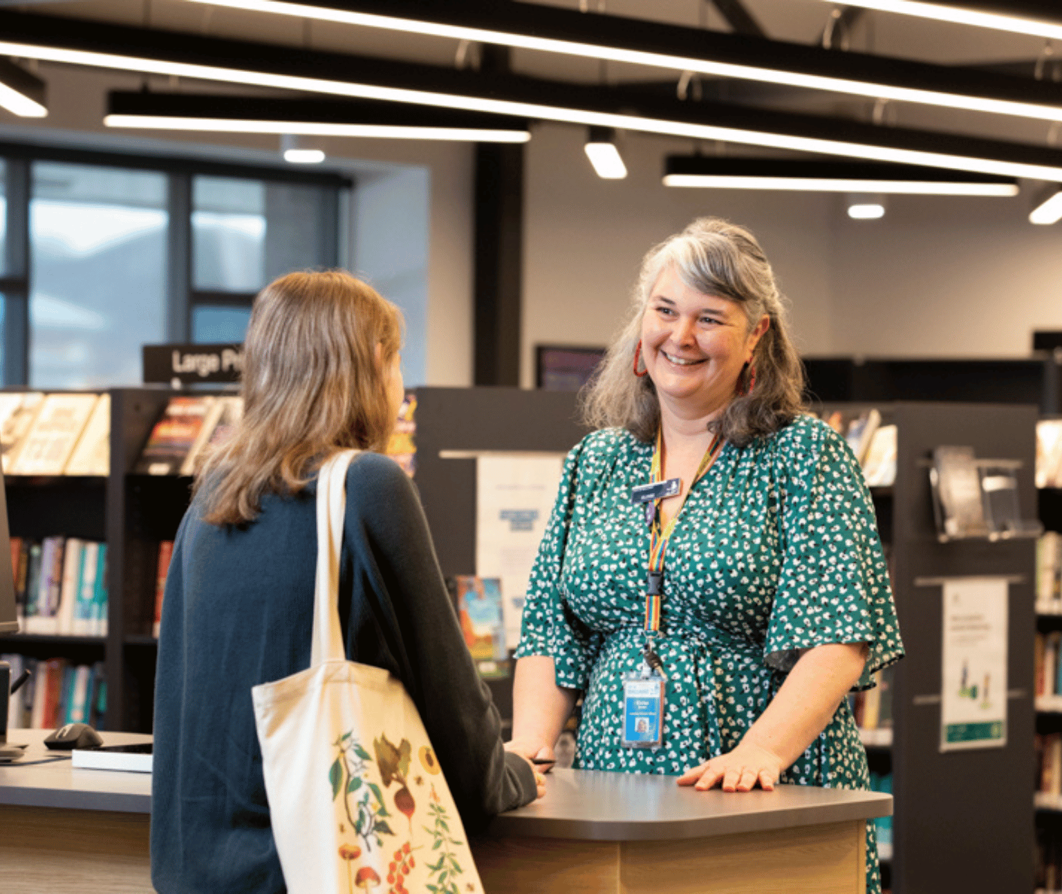 A female librarian wearing a green drees smiles at a library patron. There are shelves of books behind them.