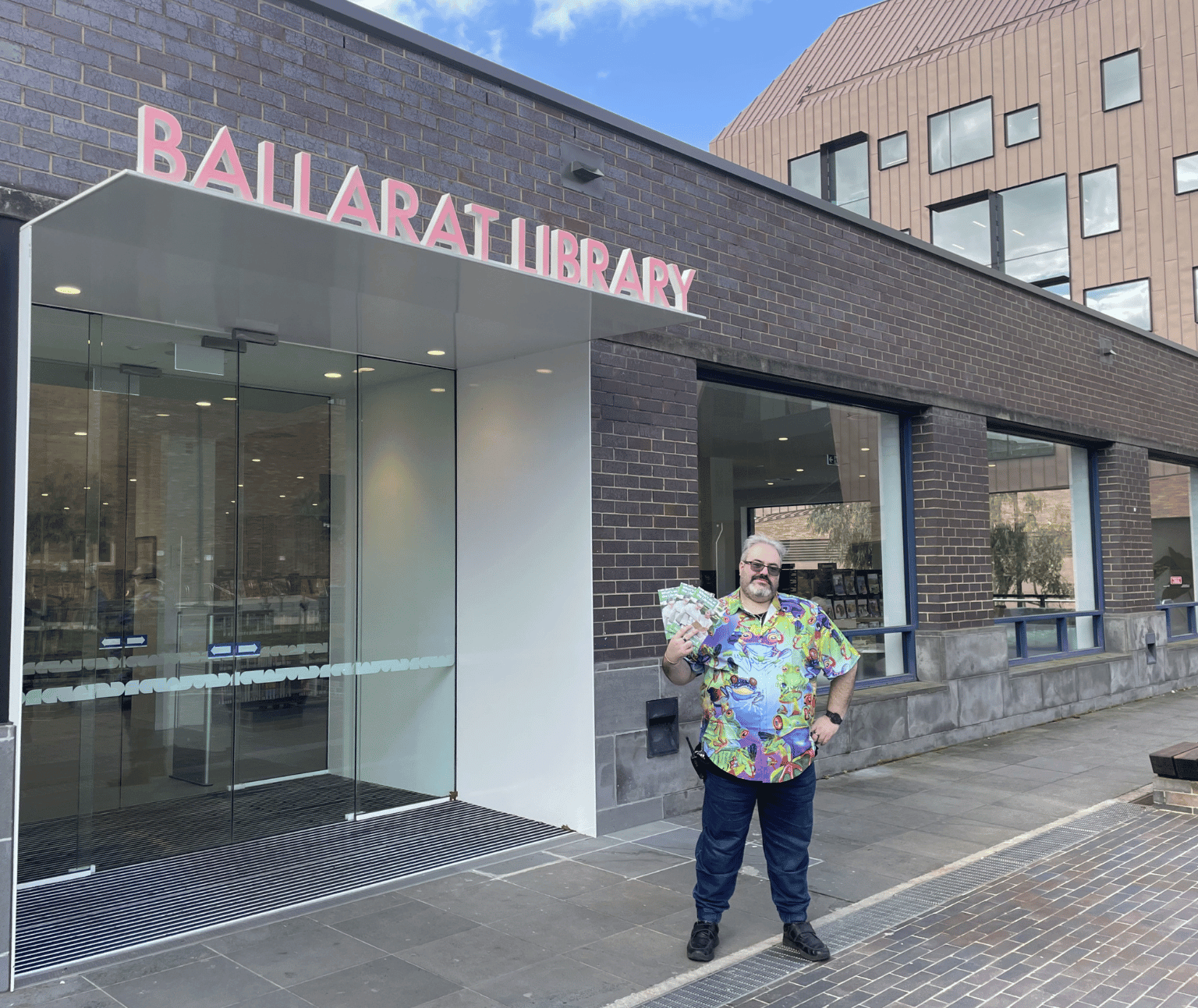A figure holds several copies of the What's On brochure in front of Ballarat Library with the sky blue and bright above them.