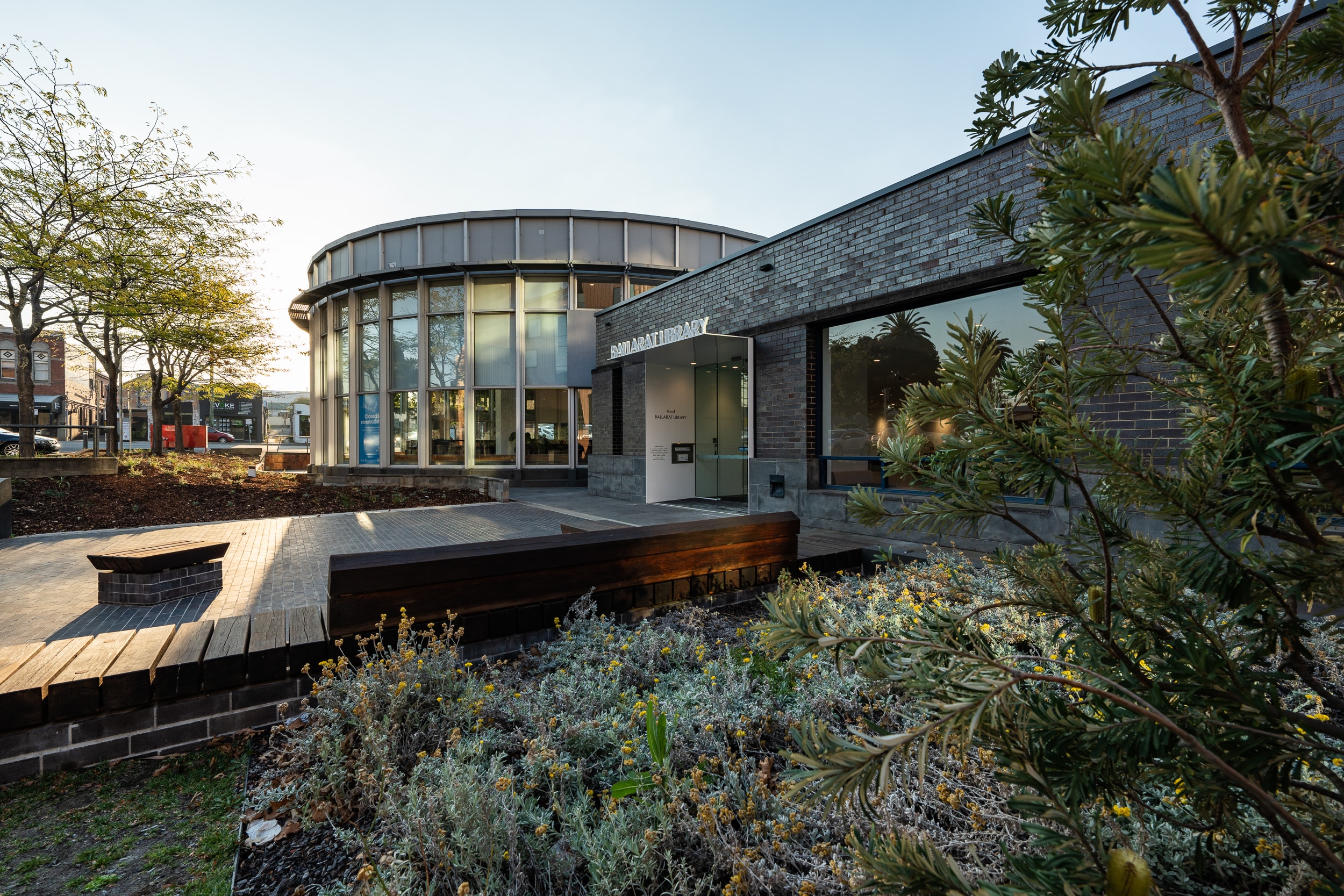 A external photo of Ballarat Library showing yellow flowers and the sun setting behind the building.