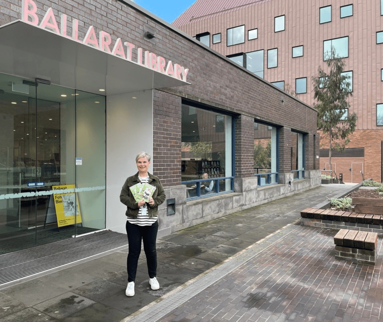 Photo of library staff member standing outside Ballarat Library holding copies of the bright green What's On in Spring brochure.