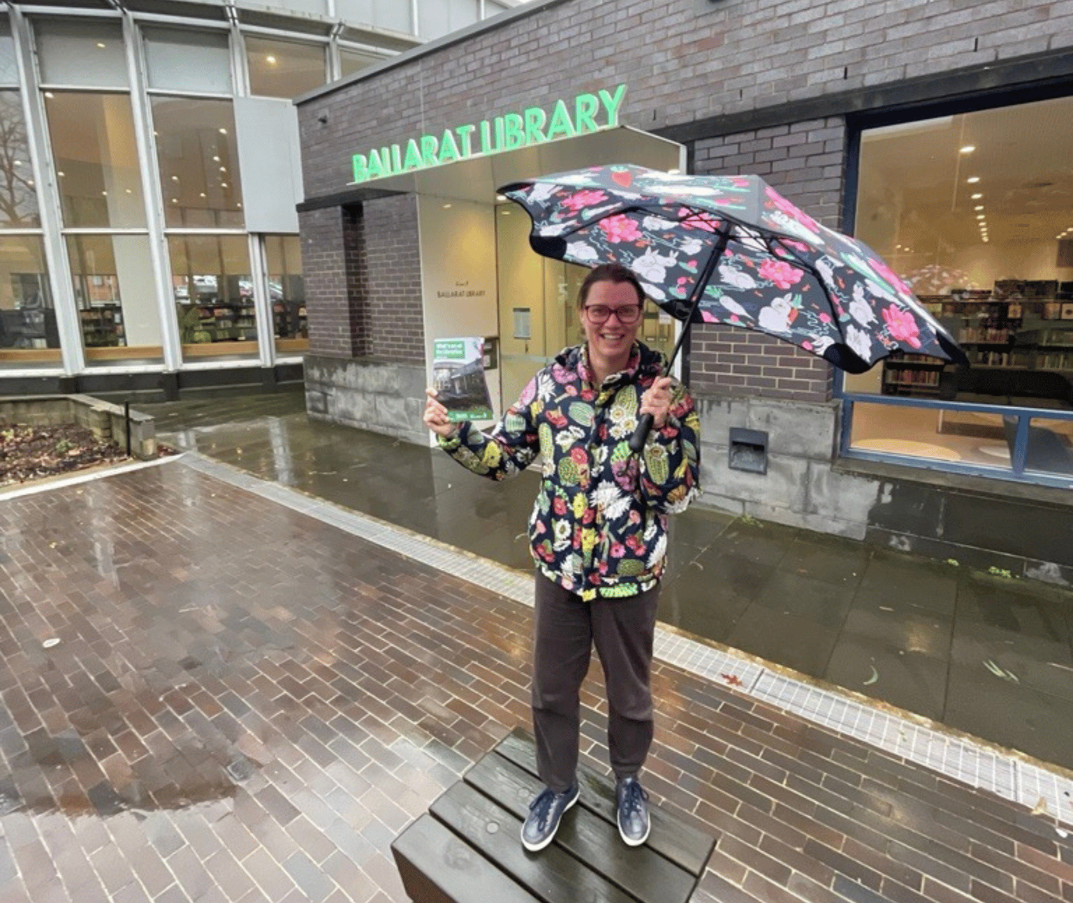 Photo of person standing in rain with umbrella and raincoat holding a copy of the Spring What's On brochure.